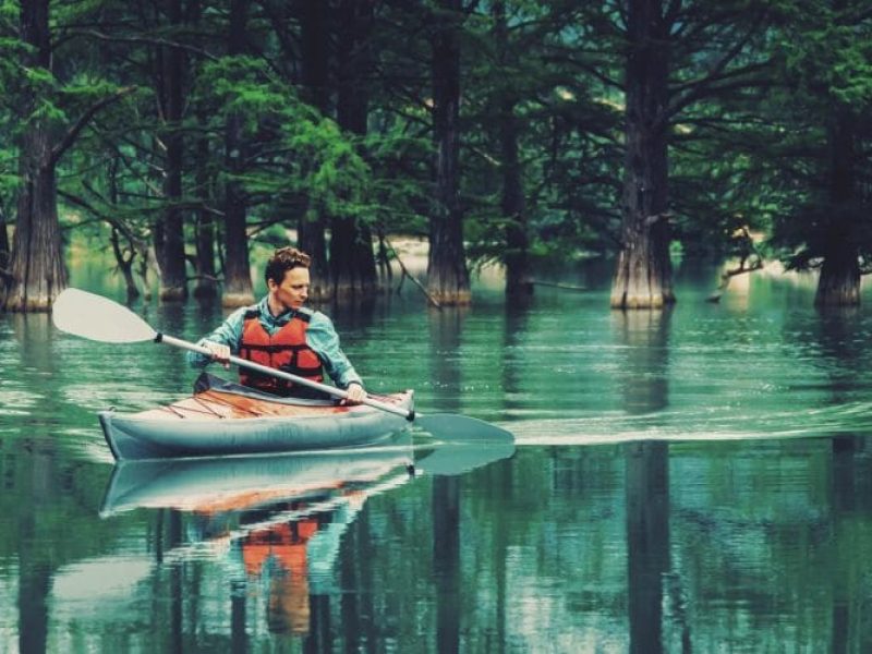 Young man paddling in kayak on lake on background of beautiful trees in summer. Toned image